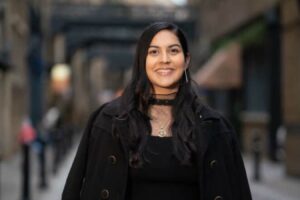 A woman in a black dress and coat stands confidently