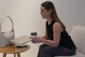 Emma sits on a chair with a coffee cup in one hand, and a laptop.