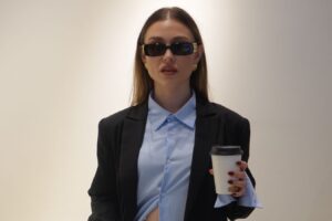 In an interview series, Emma wears a suit and sunglasses while holding a coffee cup