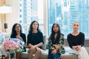 A group of women sitting on a couch, engaged in conversation, with a window providing natural light behind them
