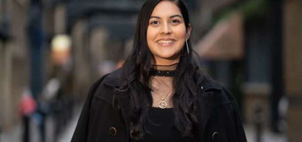 A woman in a black dress and coat stands confidently