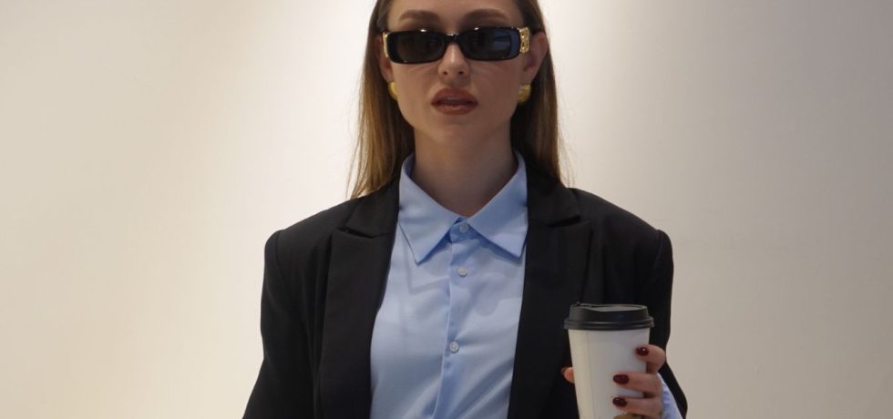 In an interview series, Emma wears a suit and sunglasses while holding a coffee cup